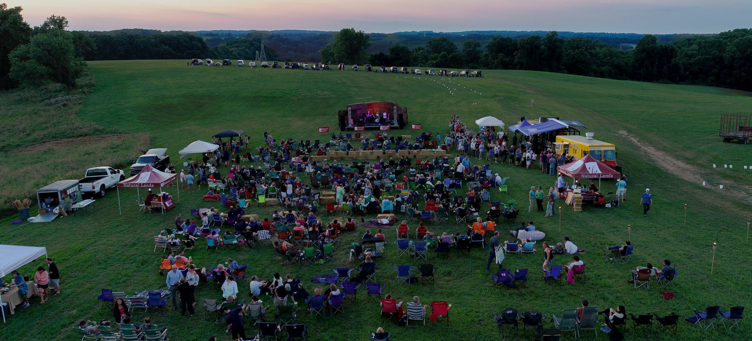 A drone shot of an outdoors concert where the audience sits on champing chairs in a green field during sunset.