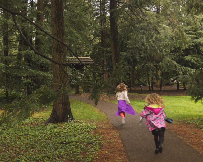 Two children with raincoats run away from the camera along a path in a wooded garden.