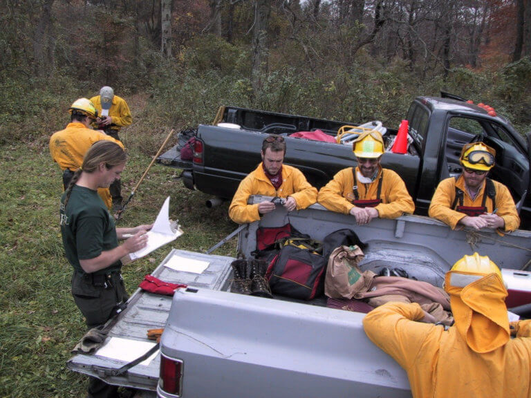 A group of people in safety fire uniforms gather around a truck in a natural landscape.