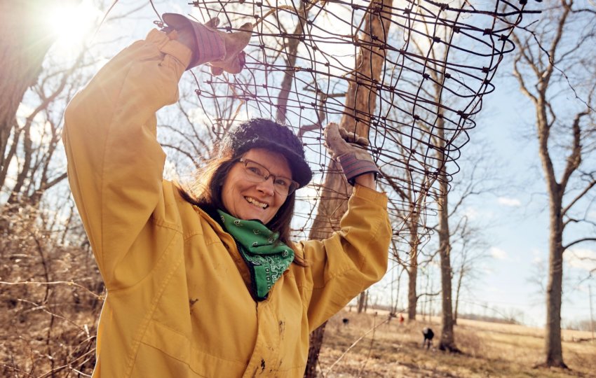 A person in a yellow jacket smiles and holds some plant fencing above their head with gloved hands as they stand in the forest.