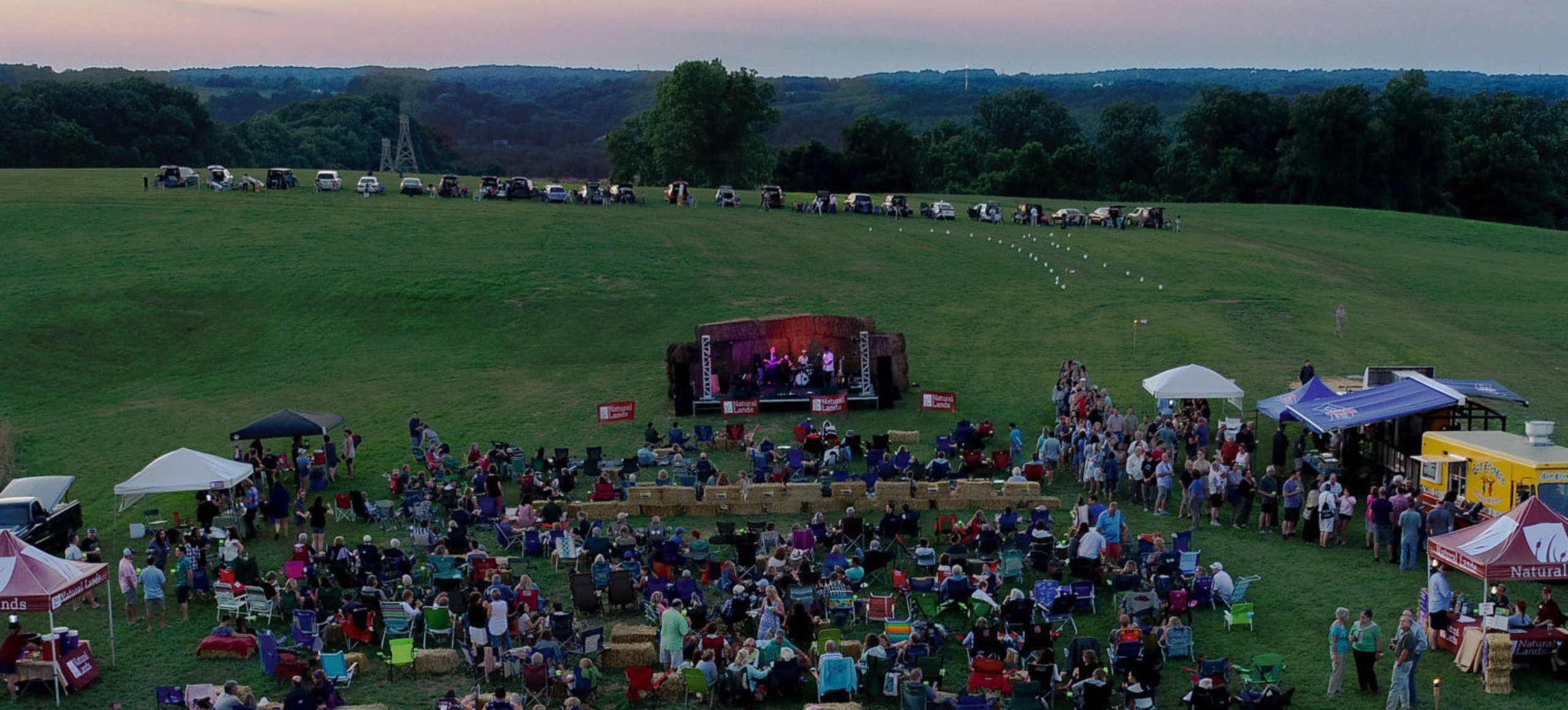 A drone shot of an outdoors concert where the audience sits on champing chairs in a green field during sunset.