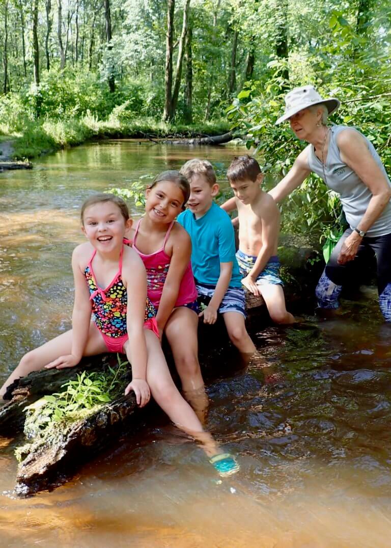 Four children sit on a log in a stream while an adult watches.