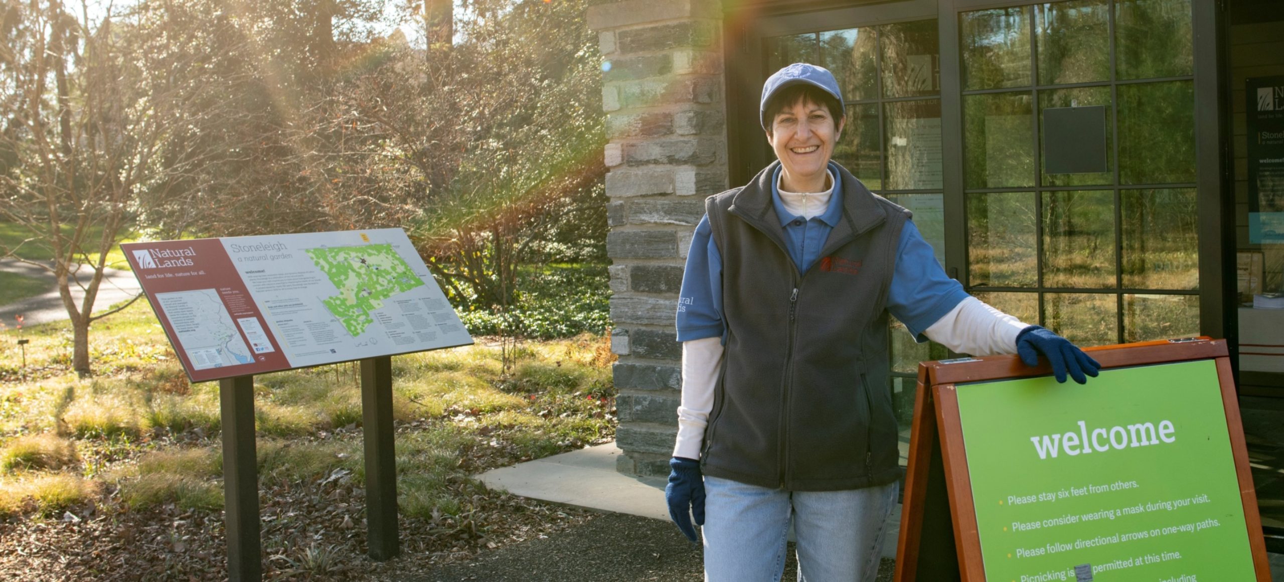 A woman in winter clothes smiles at the camera with one hand on a green welcome sign. In the backround is a map and a stone building.