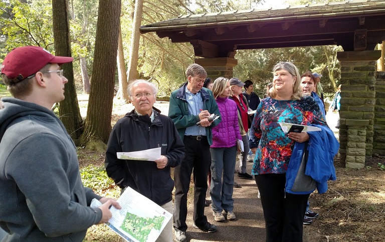 A groupd of people standing on a trail outdoors listening to a man holding a map speak.