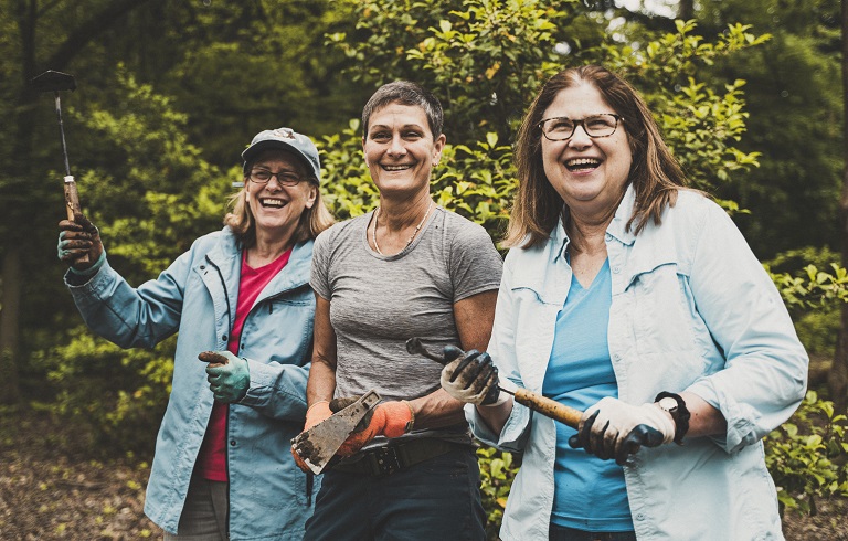 Three women holding hand tools smile at the camera outdoors.