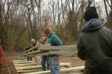 Five people lift a metal frame outdoors in a forest during winter.