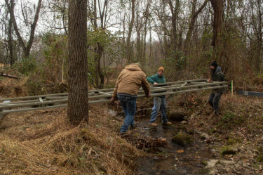 Three people carry a metal frame over a small stream outdoors in the winter.