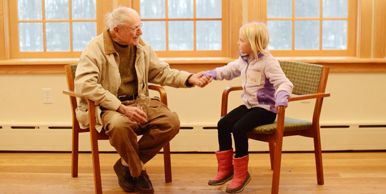 An old man and a young girl smile while holding hands sitting in wooden chairs indoors.
