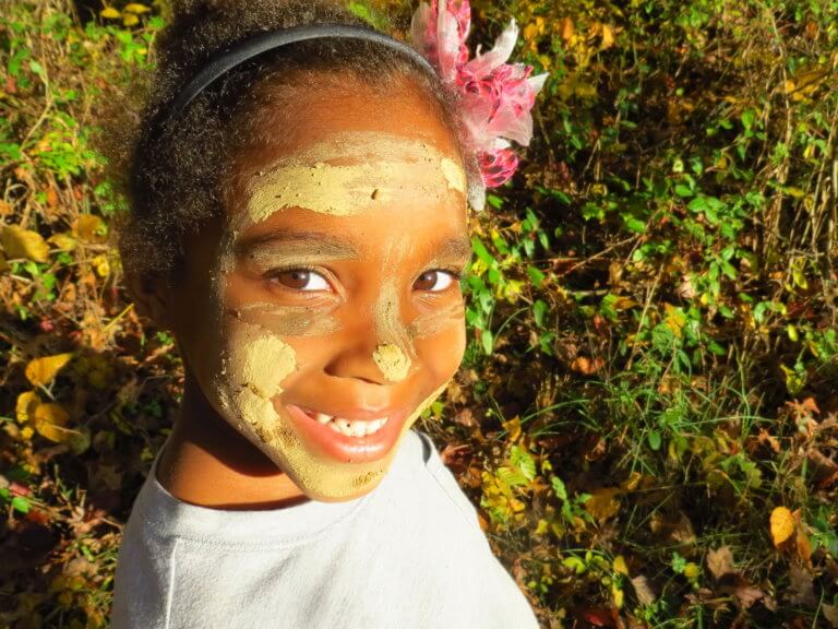 A girl with mud painted on her face smiles outdoors.