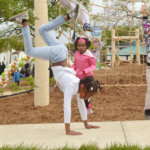 A young girl does a hand stand on a sidewalk at a playground in front of other children.