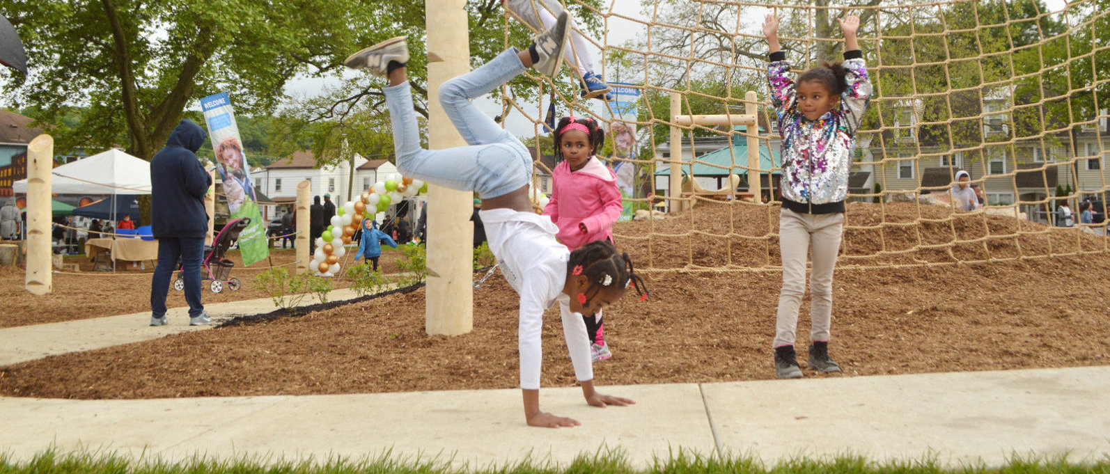 A young girl does a hand stand on a sidewalk at a playground in front of other children.
