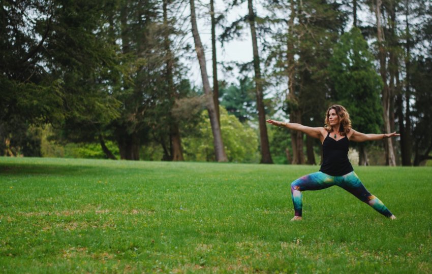 A woman does a yoga pose with outstretched arms on a grassy landscape with trees in the background.