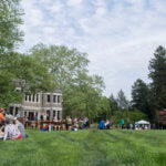 People picnic on a grassy field bordered by trees in front of a grey stone house.