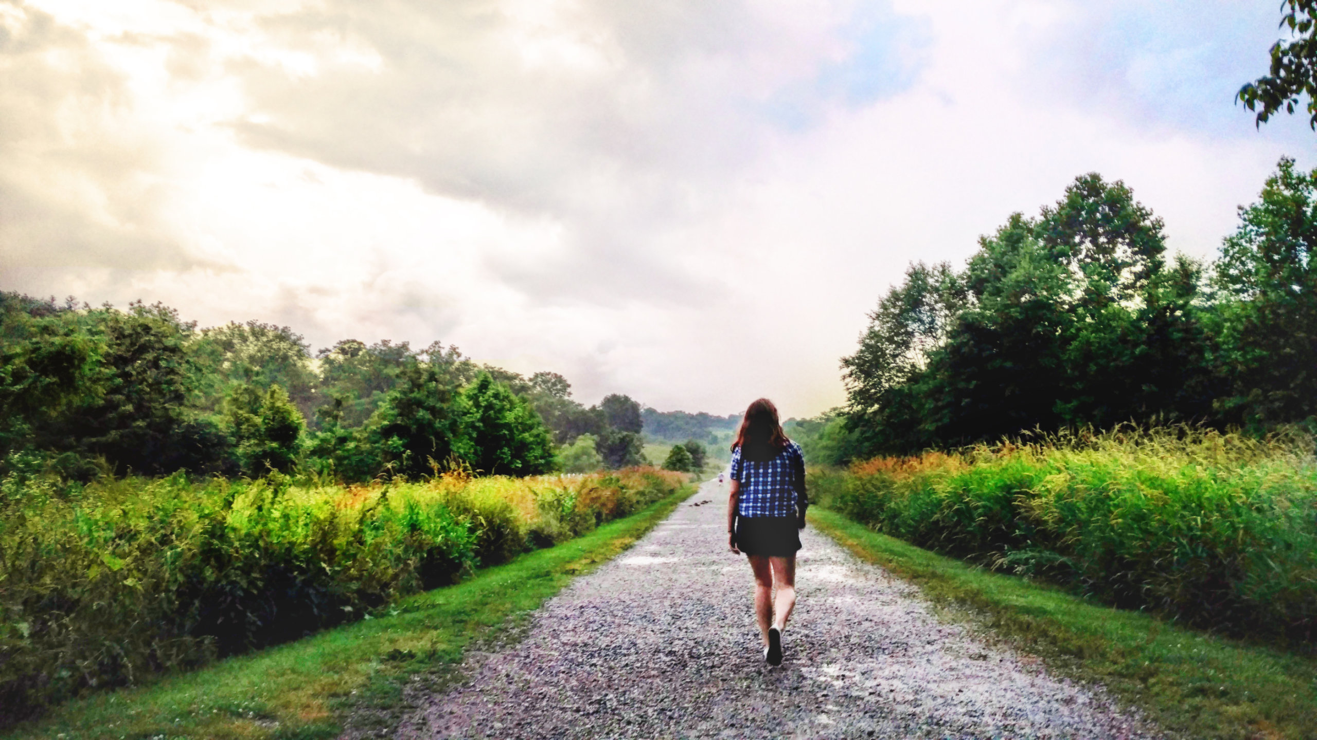 A woman walks along a long gravel path through a green natural landscape.