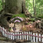 A fairy house sits in front of a tree and behind a white picket fence.