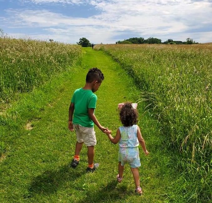 Two young children hold hands as they walk up a grass path in a green field with a blue sky.