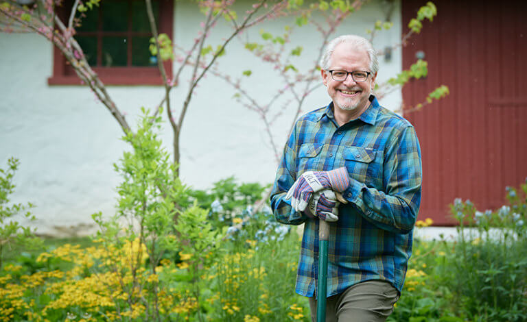 Oliver Bass smiles while wearing gardening gloves outside in front of a white and red barn.