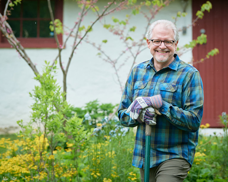 Oliver Bass smiles while wearing gardening gloves outside in front of a white and red barn.