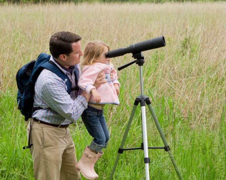 A father lifts his daughter up to look through a telescope next to a meadow.