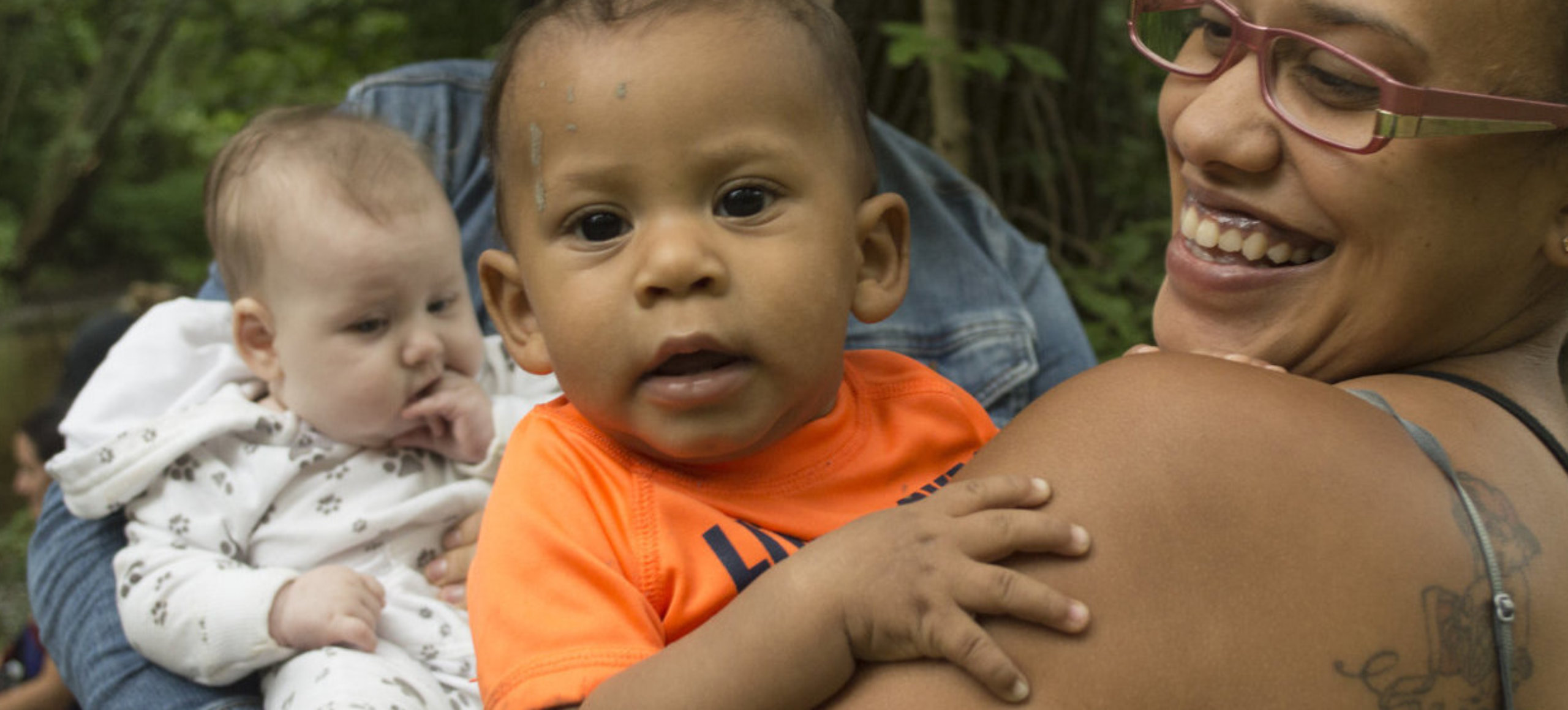A baby wearing a orange shirt looks at the camera while a woman smiles at the baby. In the backround another baby chews on their fingers.