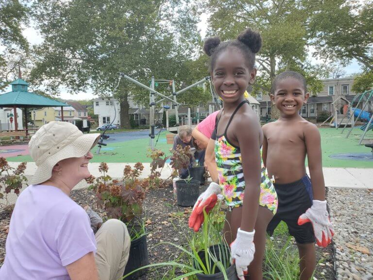 Two children wearing garden gloves smile at the camera standing in a garden at a playground. An adult sits on the ground next to them.