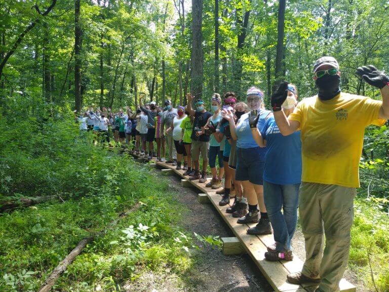 Volunteers wearing masks smile and wave while standing on a wooden boardwalk in the woods.