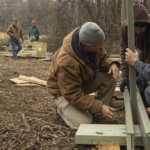 Three people in winter clothing in the foreground build a bridge out of wood and metal in front of a meadow and trees. Two people in the background look on standing in front of a pile of neatly stacked supplies.