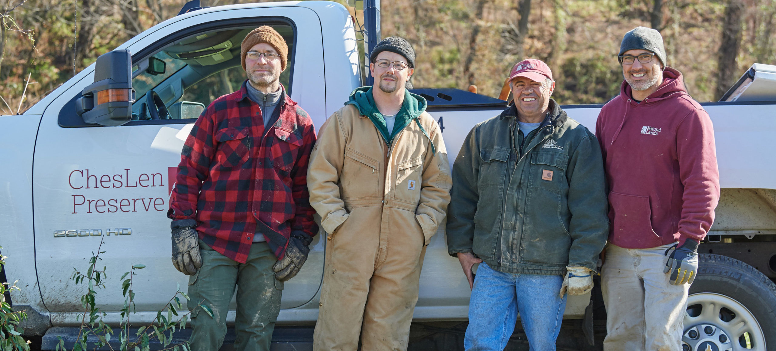 Four men in outdoor work clothes stand oudoors in front of a white truck with red letters on it that say ChesLen Preserve.