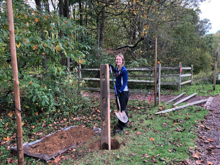 A woman holds a shovel while standing next to a hole with a fencepost in it.
