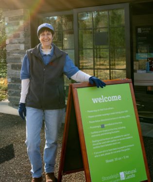 A volunteer in blue jeans, a blue shirt, grey vest and blue hat stands next to a green welcome sign in front of a stone and glass building.