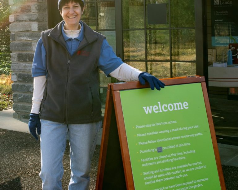 A volunteer in blue jeans, a blue shirt, grey vest and blue hat stands next to a green welcome sign in front of a stone and glass building.
