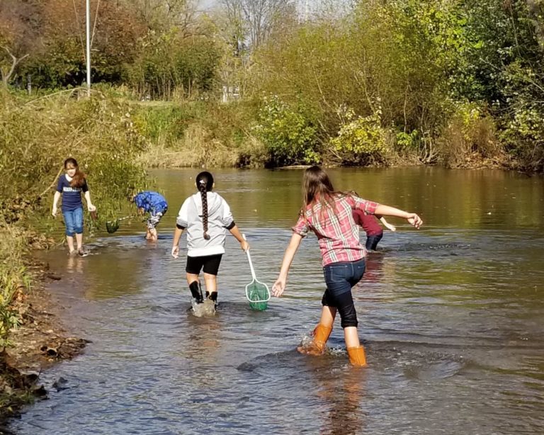Five children wade in a wide stream with boots on carrying nets for sampling aquatic life.