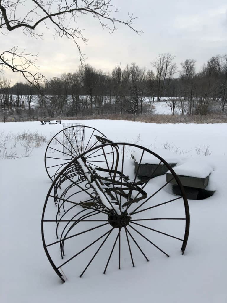 An antique farm plow rests in the foreground with its wheels in the snow. In the background there is a snowy landscape with tall trees.