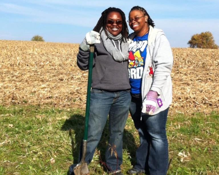 Two African-American women stand in a sunny field with shovels and work gloves.