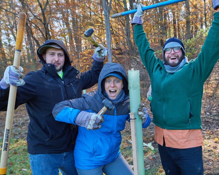 Three people outdoors wearing winter clothes pose joyfully holding hand tools. The person in the middle holds a tree tube in one hand and a hammer in the other.