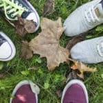 Looking down at the toes of three sets of feet in different sneakers. Green grass and fallen leaves.