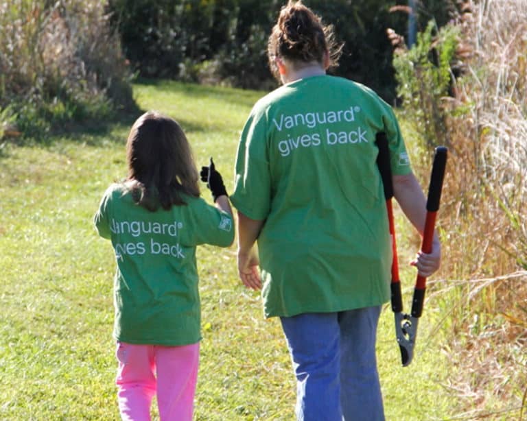 An adult and a child walk away from the camera. They are wearing work gloves and the adult is carrying loppers. They have Vanguard volunteer t-shirts on.