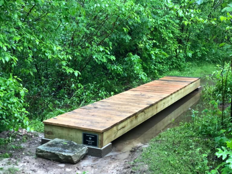 a wooden boardwalk over a calm stream with woodland greenery on both banks