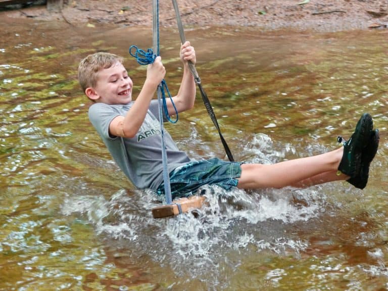 A boy smiles as he swings a wooden swing through the creek