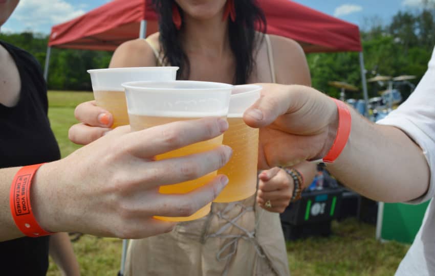 Three people clink in "cheers" with plastic cups of beer as they stand in a sunny meadow.