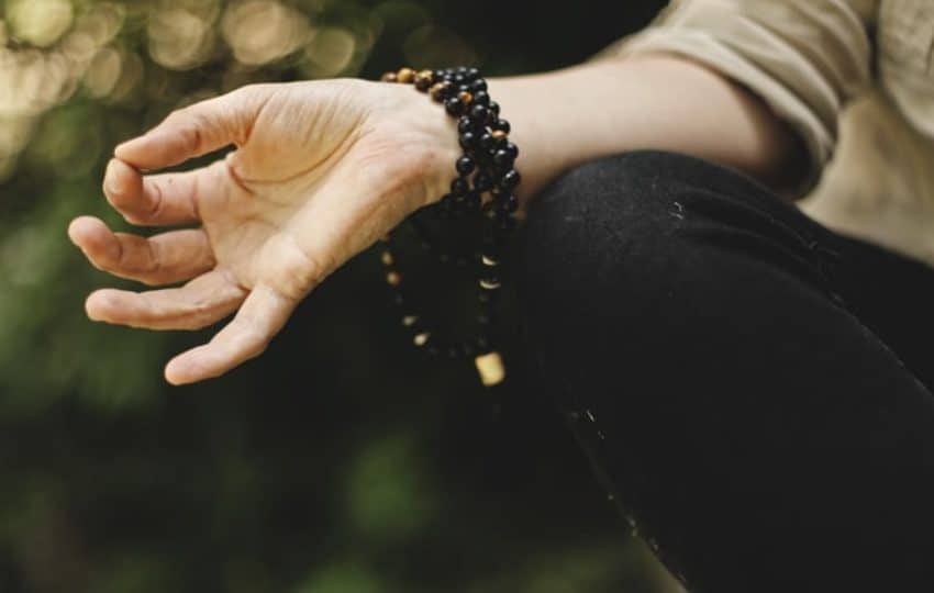 A hand in om mudra position with thumb and forefinger touching, arm with prayer beads resting on crossed legs outside