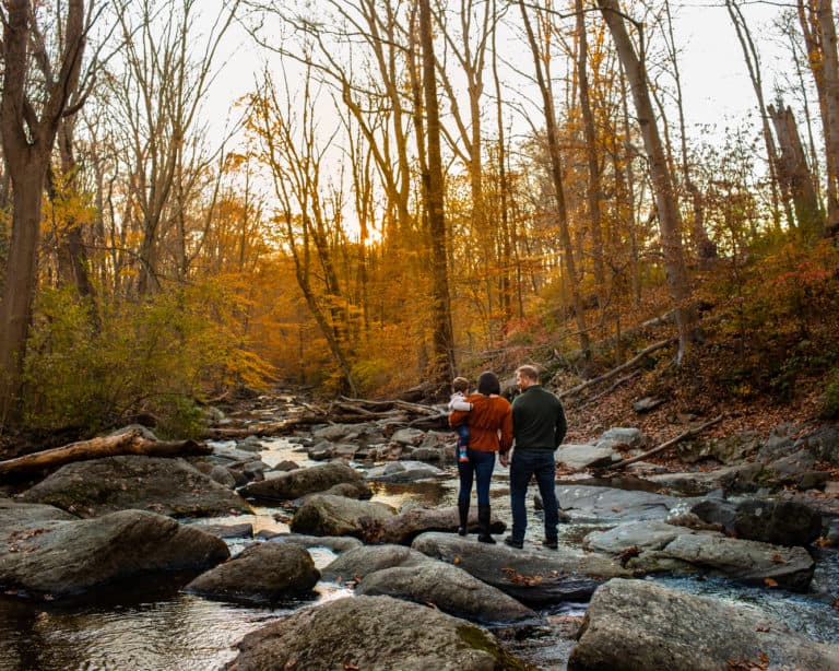 A woman holding a baby holds hands with a man standing on a rock in the middle of a stream in an autumn forest.