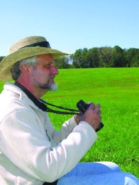 A man in a white shirt and hat looks across a meadow with binoculars in his hands.