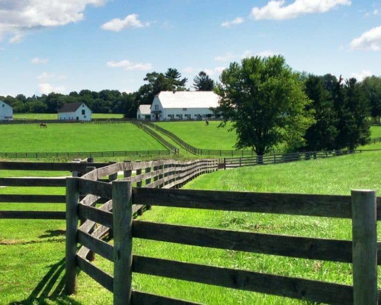 A zig-zagging wooden fence through green farm fields with a barn and blue skies.