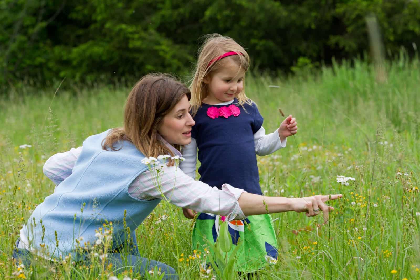 A mother and a young girl stand in a meadow with wildflowers. The mother is pointing to a white bloom and the daughter is looking at it.