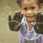 a smiling child with dark brown eyes and hair holds muddy hands up at the camera with a body of water behind them.