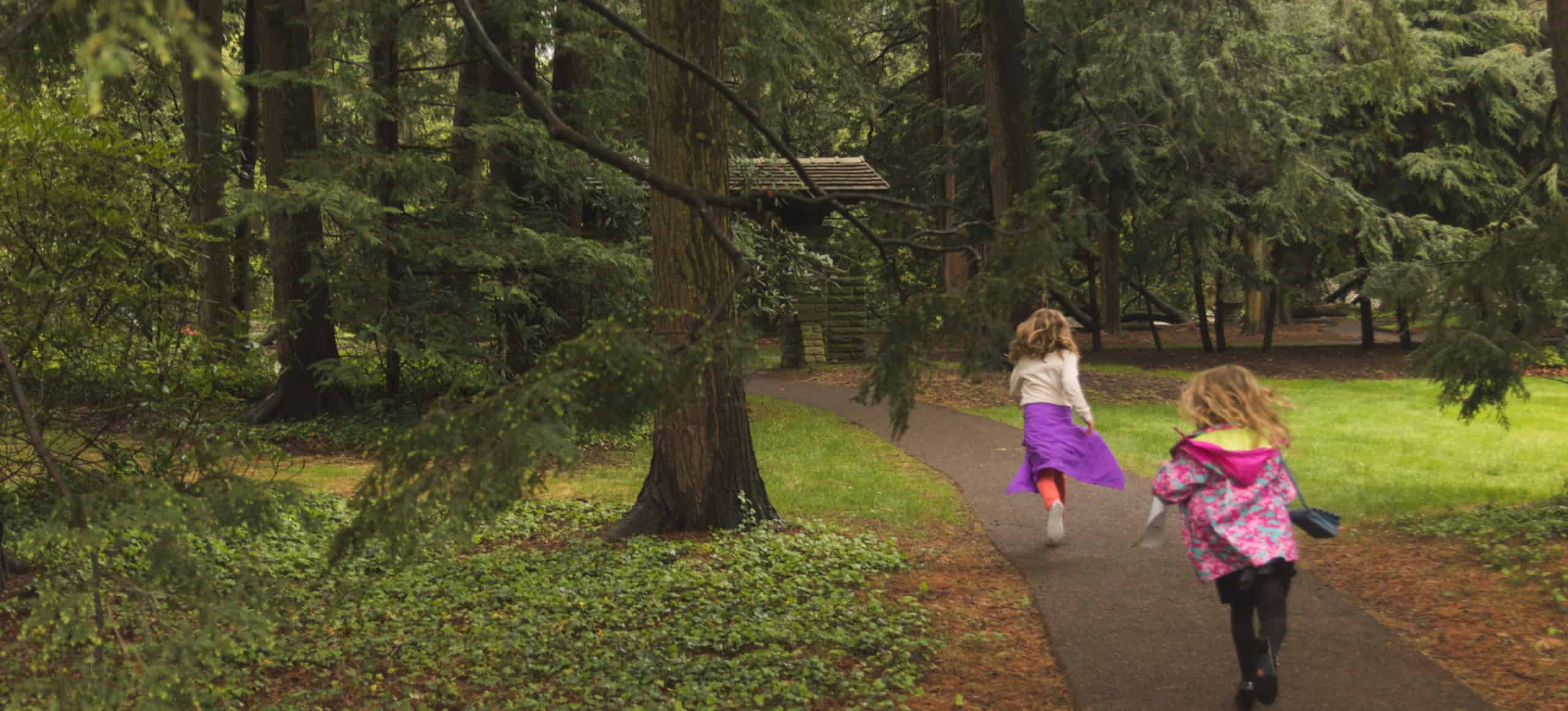 Two children run along a path through a natural setting with tall trees.
