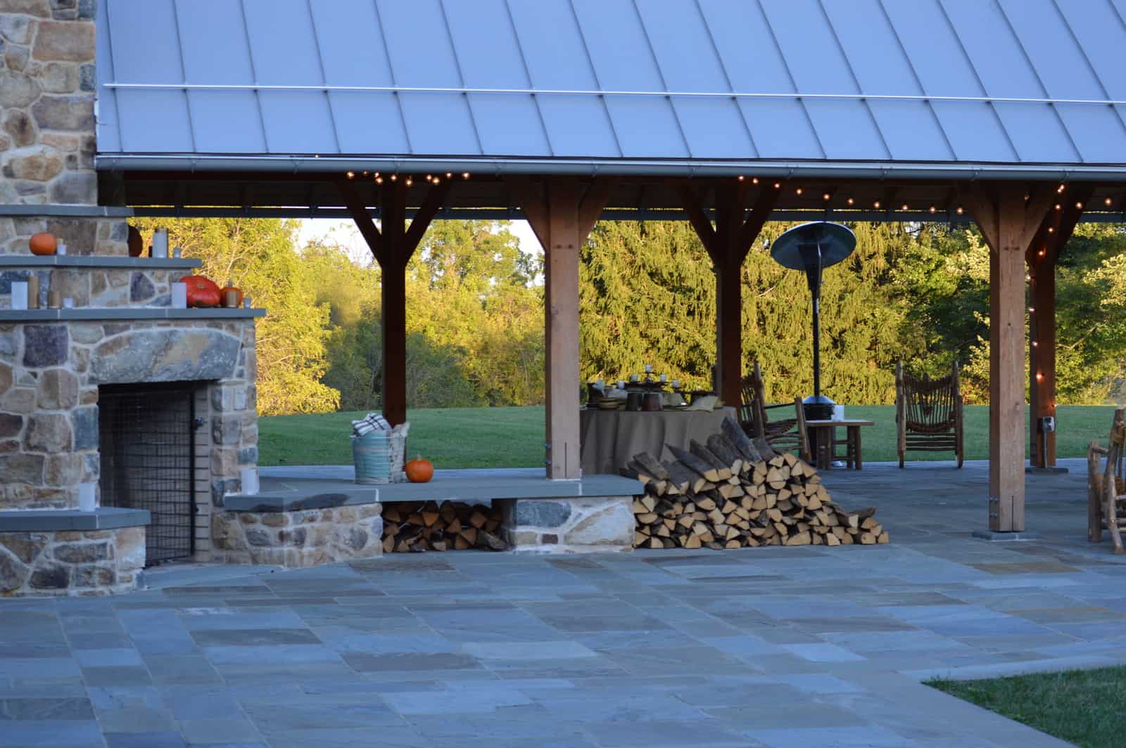 A stack of split firewood next to an outdoor stone fireplace near an open-air pavilion.