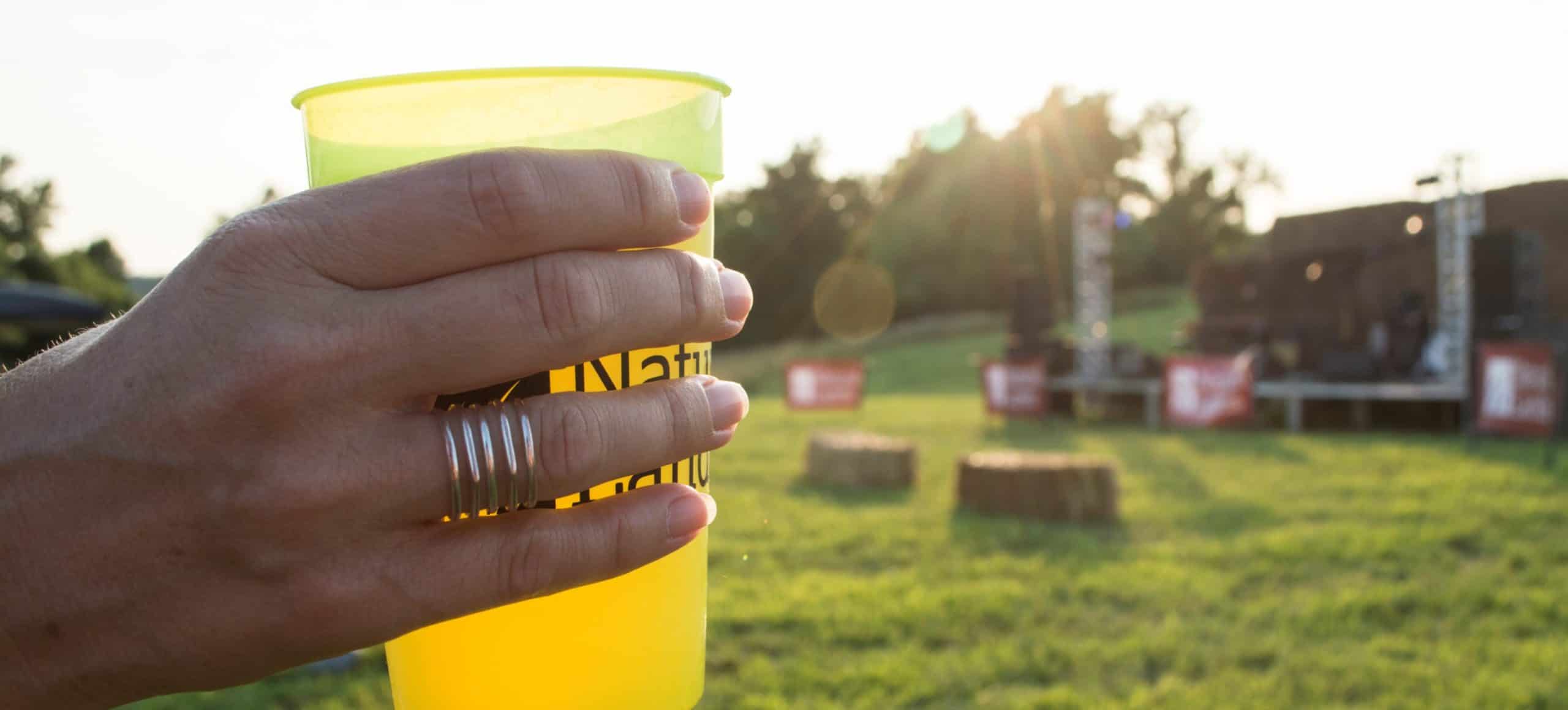 Close up of a hand holding a pint of beer with the sun and grassy space behind