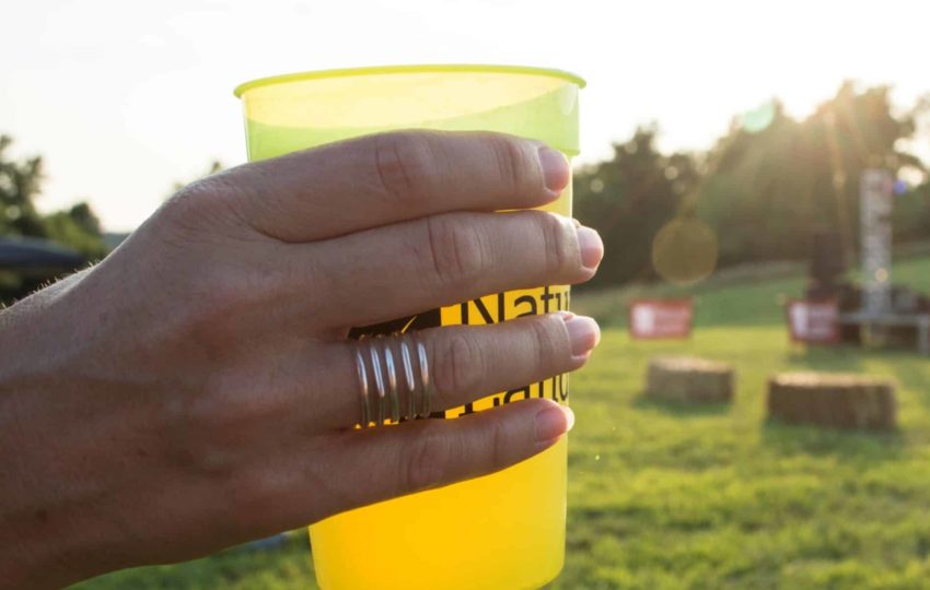 Close up of a hand holding a pint of beer with the sun and grassy space behind